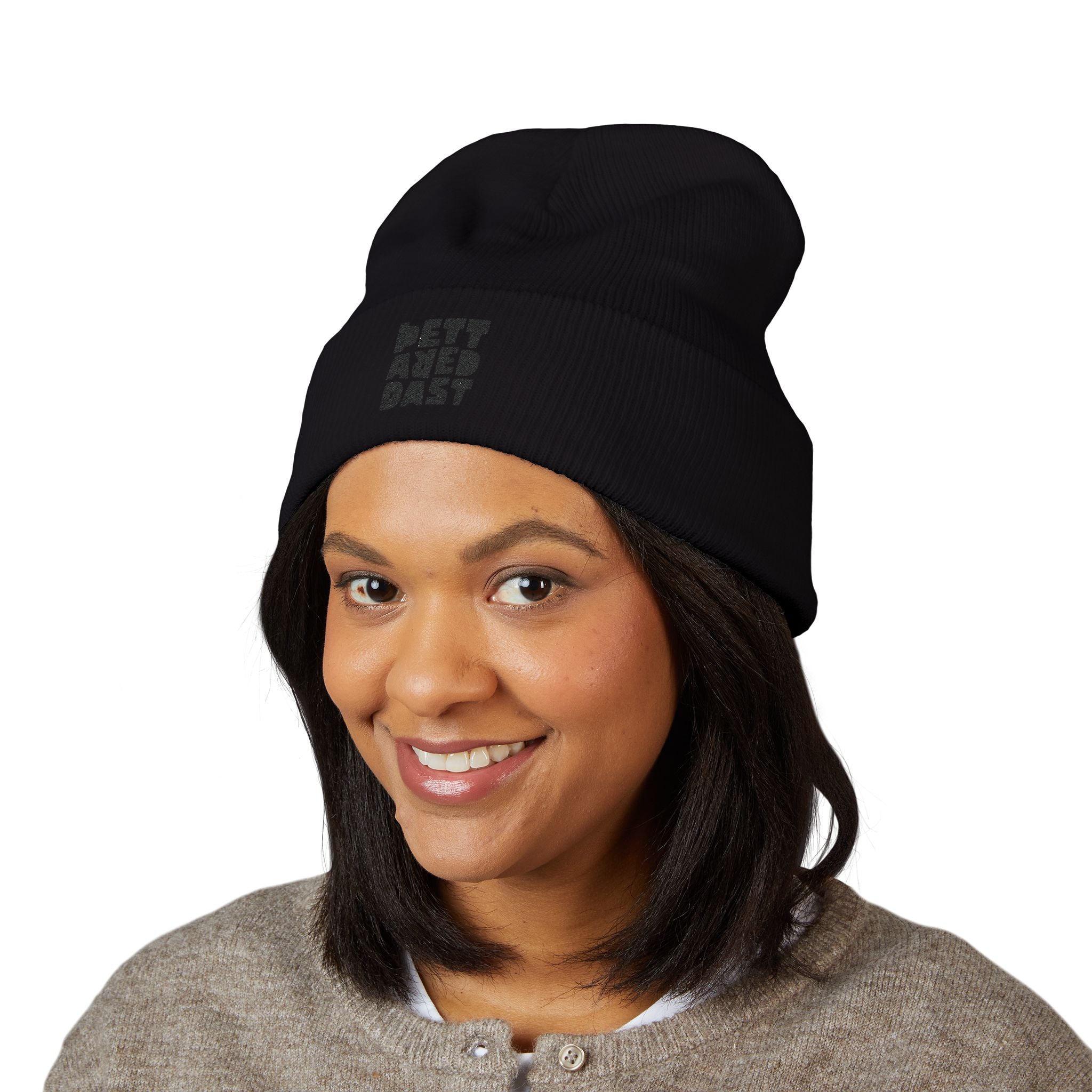 A woman with shoulder-length dark hair smiles against a plain white background, wearing Printify’s Embroidered Cuffed Beanie—Subtle 'Pet Area Baby' black knit hat and a beige sweater.