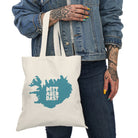 A person in a denim jacket holds the Printify Natural Tote Bag made from sustainable cotton, featuring a blue map of Iceland and the text “Þetta Ætla Ég Að Dasta.”.