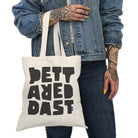 A person in a denim jacket and jeans holds the Tote Bag Natural by Printify, a sustainable cotton bag with bold abstract black text, and sports colorful nail polish.