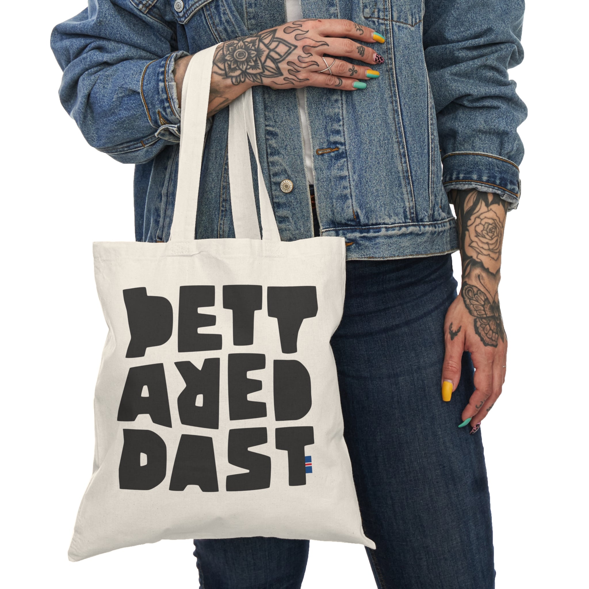 A person with tattooed hands in a denim jacket holds Printify's Copy of Natural Tote Bag, custom printed with bold black block letters spelling "DETT AR E DAST.